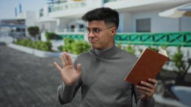 Şehir dışında kitap okuyan genç bir adam. Düşünceli bir ifadeyle gri balıkçı yaka kazak ve bulanık şehir zeminine karşı gözlük takıyor.