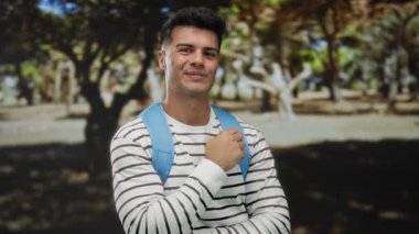 Güneşli bir günde açık hava parkında sırt çantasıyla gülümseyen genç bir adam..