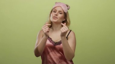 Pembe gecelikli genç sarışın kadın kalp hareketi yapıyor. Sarı arka plan karşısında neşeli bir uyku maskesi takıyor. Sevgi ve rahatlama hissi veriyor..