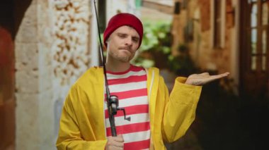 Sarı ceketli ve kırmızı bereli genç adam elinde olta kamışı tutarken tuhaf bir şehir arka planında, düşünceli ve rahat bir havayı çağrıştırıyor..