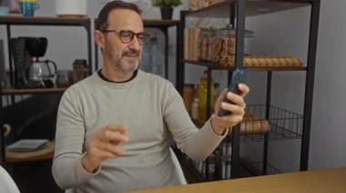 Adam modern ofiste raflarda çeşitli eşyalar sergilenirken selfie çekiyor, akıllı telefonu tutarken gülümsüyor, iş ortamında dostane ve profesyonel bir atmosfer yaratıyor..