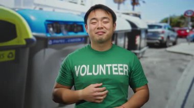Gönüllü gömleği giyen genç adam, şehir sokaklarındaki geri dönüşüm kutularının yanında güvenle duruyor. Çevresel sorumluluğu ve toplumsal bağlılığı teşvik ediyor..