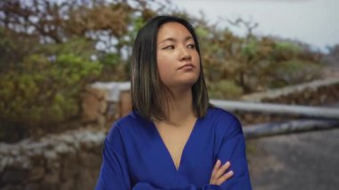 Mavi elbiseli kadın, gün ışığında bulanık doğal geçmişi olan parkta çapraz kollarıyla duruyor. Açık havada sakin kalmayı öneriyor..