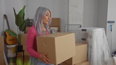 Gri saçlı yaşlı bir kadın yeni evinde kutu taşıyor. Etrafı kutular ve paketsiz eşyalarla çevrili. Bu da sıcak bir oturma odasında taşınma günü olduğunu gösteriyor..