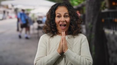 Kıvırcık saçlı bir kadın şehir sokaklarında heyecan ifade ediyor, süveter giyiyor ve neşe sergiliyor, açık hava kentsel ortamında yumuşak odaklı arka planda olgun bir güzellik sergiliyor..