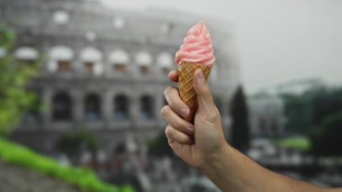 Roma Kolezyumu 'nun önünde pembe bir dondurma külahı tutarken arka planda bulanıklık vardı. Roma' da güneşli bir açık hava sahnesini beyaz bir adamın belirgin bir şekilde öne çıkan eliyle çekiyordu..