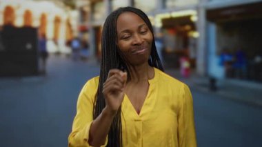 Sarı bluzlu, kıvırcık saçlı bir kadın şehir sokaklarında kendine güvenerek duruyor ve çarpıcı varlığıyla şehrin atmosferini yakalıyor..