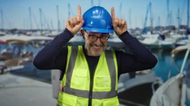 Güvenlik teçhizatı ve kasket takan bir adam huzurlu sahil limanındaki teknelerle eğlenceli jestler yapıyor..