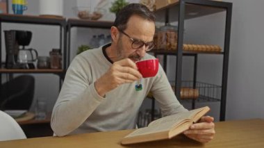 Elinde kırmızı bardakla kitap okuyan olgun bir adam. Etrafı raflar ve kahve ekipmanlarıyla çevrili..