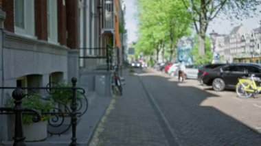 Amsterdam 'da insanlar, bisikletler ve binalarla dolu bulanık bir sokak manzarası. Canlı yeşillik ve yol kenarında park halindeki arabalarla klasik bir şehir atmosferi yaratıyor..