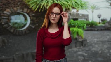 Şehir caddesinde yeşil bitkilerle gözlük takan bir kadın kızıl saçlarını ve kırmızı süveterini gösteriyor..