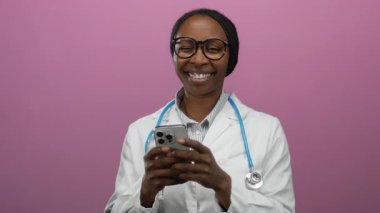 Doktor gülümserken pembe arka planda cep telefonu kullanıyor, steteskop takıyor, büyük gözlük takıyor, beyaz önlük takıyor, profesyonel iletişim ve mutluluk taşıyor.