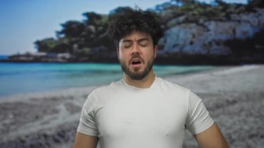 Güneşli bir sahilde hapşıran sakallı genç İspanyol adam, sakin okyanus dalgaları ve kayalık uçurumlarla çevrili serin deniz meltemini hissediyor..