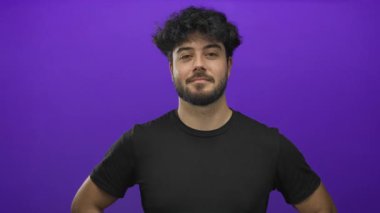 Mor arkaplan önünde siyah tişörtlü, sakallı genç adam stüdyo ortamında özgüvenle gülümsüyor, gündelik tarzını ve pozitif tutumunu sergiliyor..