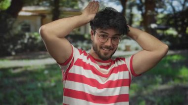 Gözlüklü, yeşil bir parkta kulak hareketi yapan, neşeli çizgili bir tişörtle güneşli ağaçların altında gülümseyen İspanyol bir genç..