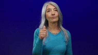 İzole edilmiş mavi arka planda gri saçlı yaşlı kadın turkuaz süveter giyiyor ve tuhaf bir jest yapıyor.