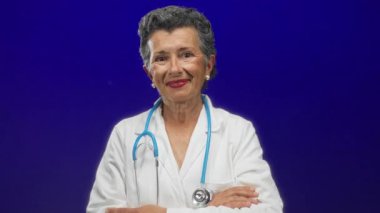 Steteskoplu beyaz önlüklü kıdemli bayan doktor, mavi arka planda, profesyonellik ve samimiyet yayıyor, sağlık teması için uygun..