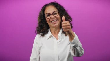Kıvırcık saçlarıyla gülümseyen bir kadın canlı mor arka plana başparmak işareti vererek pozitiflik ve başarı gösteriyor..