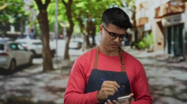 Kırmızı kazak ve kot önlük giyen İspanyol genç adam ağaçlarla ve park halindeki arabalarla dolu bir sokakta kalemle notlar alıyor.