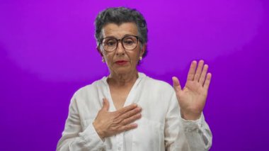 Gri saçlı yaşlı kadın canlı mor bir arka plana karşı yemin ediyor, gözlük ve beyaz gömlek takıyor, samimiyet ve bağlılık taşıyor..