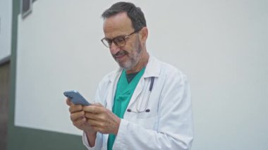 Dışarıda duran doktor, beyaz önlüklü ve steteskoplu akıllı telefona bakarken gülümsüyor..