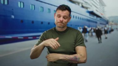 Deniz kıyısındaki bir limanda, arkasında büyük bir yolcu gemisi olan genç adam, açık havada rahat bir şekilde konuşurken merak ve merak gösteriyor..