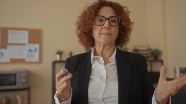 Ofis ortamında konuşan, kıvırcık esmer saçlı, profesyonel giyimli ve İspanyol kökenli orta yaşlı profilini vurgulayan bir kadın..