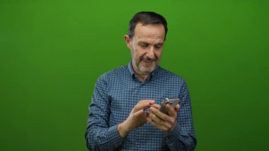 Yeşil arka plan önünde akıllı telefon kullanan gülümseyen adam kapalı alanda izole edilmiş mutluluk ve modern teknolojiyi gösteriyor.