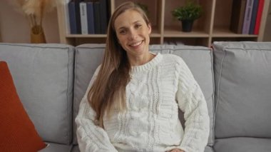 Modern oturma odasındaki rahat beyaz kazaklı, uzun saçlı kanepede gülümseyen kadın kitap rafları ve yastıklarla iç mekanda sıcak ve davetkar bir atmosfer yaratıyor..