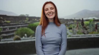 Açık hava parkında gülümseyen bir kadın kızıl saçlı ve mavi kazaklı doğal güzelliğini sergiliyor. Etrafı yeşillik ve taş oluşumlarla çevrili..