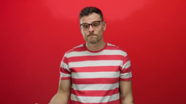 Gözlüklü genç adam canlı kırmızı bir arkaplanda poz veriyor, çizgili bir gömlek giyiyor, kendine güvenerek el kol hareketi yapıyor, günlük tarzını ve gençlik enerjisini somutlaştırıyor..