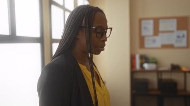 Ofisteki gözlüklü bir kadın stresli görünüyor, bir iş ortamında bir pencere ve kitaplığın yanında duran iş zorluklarını düşünüyor.