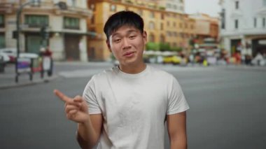 Şehir caddesinde ihtiyatlı bir jest yapan genç adam, arka planda binalar ve bulanık yayalarla canlı şehir yaşamını ve kültürel çeşitliliği vurguluyor..