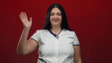 Uzun saçlı, gülümseyen ve kırmızı arka planda el sallayan, kendine güveni ve pozitif enerjisini canlı bir ortamda somutlaştıran bir kadın..