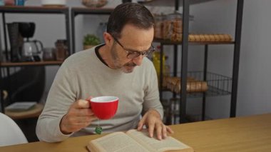 Ofiste kitap okuyan olgun bir adam düşünceli bir iş ortamı için etrafı raflarla ve mutfak gereçleriyle çevrili kırmızı bardakla çay poşeti tutarken..