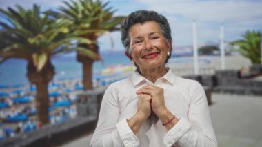 Yaşlı bir kadın, deniz kenarında palmiye ağaçları ve arka planda kumlu bir plajla neşeyle gülümseyen deniz kenarında huzurlu bir anı yakalıyor..