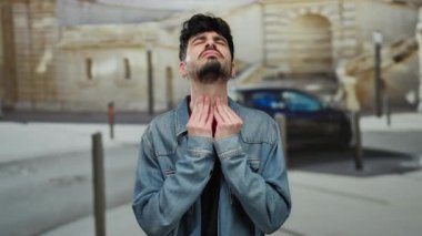 Açık şehir ortamında boğazını tutan kot giyen genç İspanyol bir adam kasabanın arka planına karşı rahatsızlık gösteriyordu..