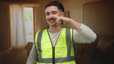 Otelin içinde yansıtıcı yelek giyen genç İspanyol bir adam, burnuna işaret ederek, pencerelerden içeri güneş ışığı süzülen sıradan ve eğlenceli bir atmosfer yaratıyor..