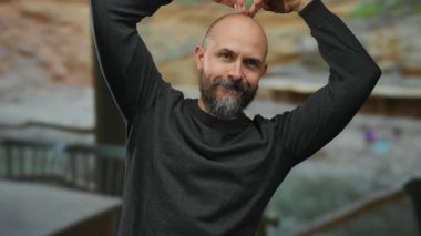 Kel ve sakallı bir adam, arka planda okyanusla birlikte sahilde gülümsüyor, açık havada günlük giysiler giyiyor ve güneşli bir günün tadını çıkarıyor..