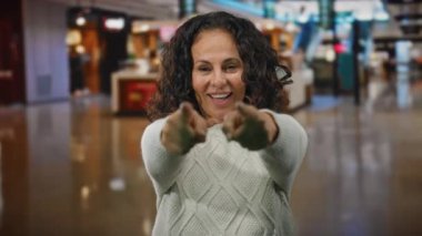 Alışveriş merkezinde kıvırcık saçlı bir kadın, açık bir gülümsemeyle davet ediyor, beyaz bir kazak giyiyor, alışveriş ortamında sıcaklığı ve arkadaşlığı somutlaştırıyor..