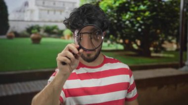 Sakallı genç adam açık havada güneşli bir arka planda büyüteç tutuyor, merakla araştırıyor ve gözlemliyor, parlak bir günde çizgili bir gömlek giyiyor..