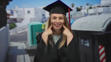 Genç bir kadın, açık şehir ortamında elde ettiği başarıyı gösteren neşeli bir ifadeyle, kep ve cübbe giyerek şehir caddesinde mezuniyetini kutluyor..