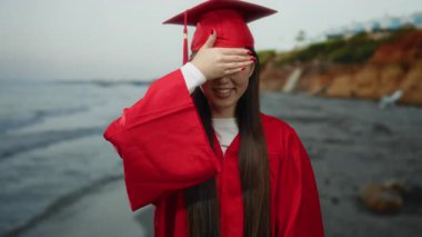 Sahilde mezuniyeti kutlayan kırmızı şapkalı ve üzerinde okyanus dalgaları olan cüppeli bir kadın.
