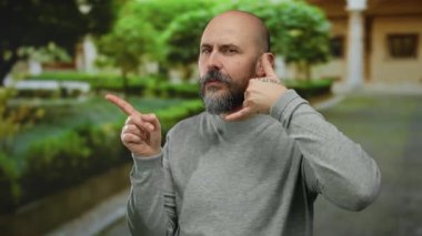 Şehir sokağında yeşil arka planda, kel, sakallı, gri kazak giyen, kendine güvenen, yakışıklı bir yüz ifadesi olan, açık hava manzaralı, şehir manzaralı bir adam..