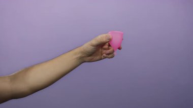 Mor arka planda pembe regl bardağı tutan adam, hijyen ve cinsiyet ayrımcılığını vurguluyor..