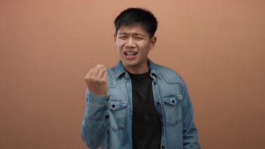 İzole edilmiş bir kahverengi duvar arkaplanına el hareketleriyle şüphecilik gösteren genç bir adam, günlük bir kot ceket giyerken belirsizlik ve tefekkür taşıyor.