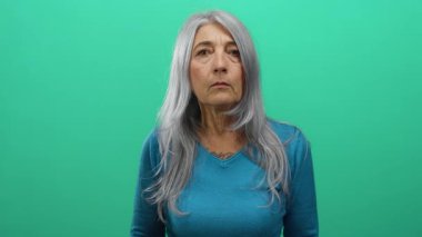 Son sınıf öğrencisi, gri saçlı, mavi kazaklı, yeşil arka plan duvarına yaslanmış sakin ve dingin bir kadın..