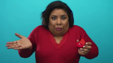 Mavi arka planda sürpriz bir ifadeyle yüzük kutusu tutan bir kadın kırmızı bir kazak giyiyor ve beklenmedik tekliflerle ilgili duygu ve jestler sergiliyor..