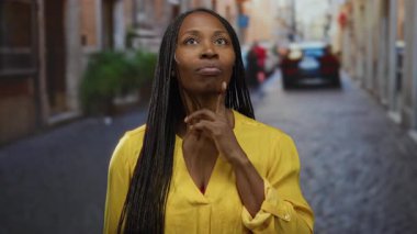 Sarı tişörtlü bir kadın şehir caddesinde çenesinde parmak gezdiriyor..