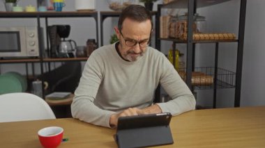 Ofisteki olgun İspanyol bir adam, tablet kullanarak video görüşmesi yapıyor. Etrafı modern mutfak malzemeleri ve kırmızı bardakla çevrili. Odaklanmış ve profesyonel görünüyor..